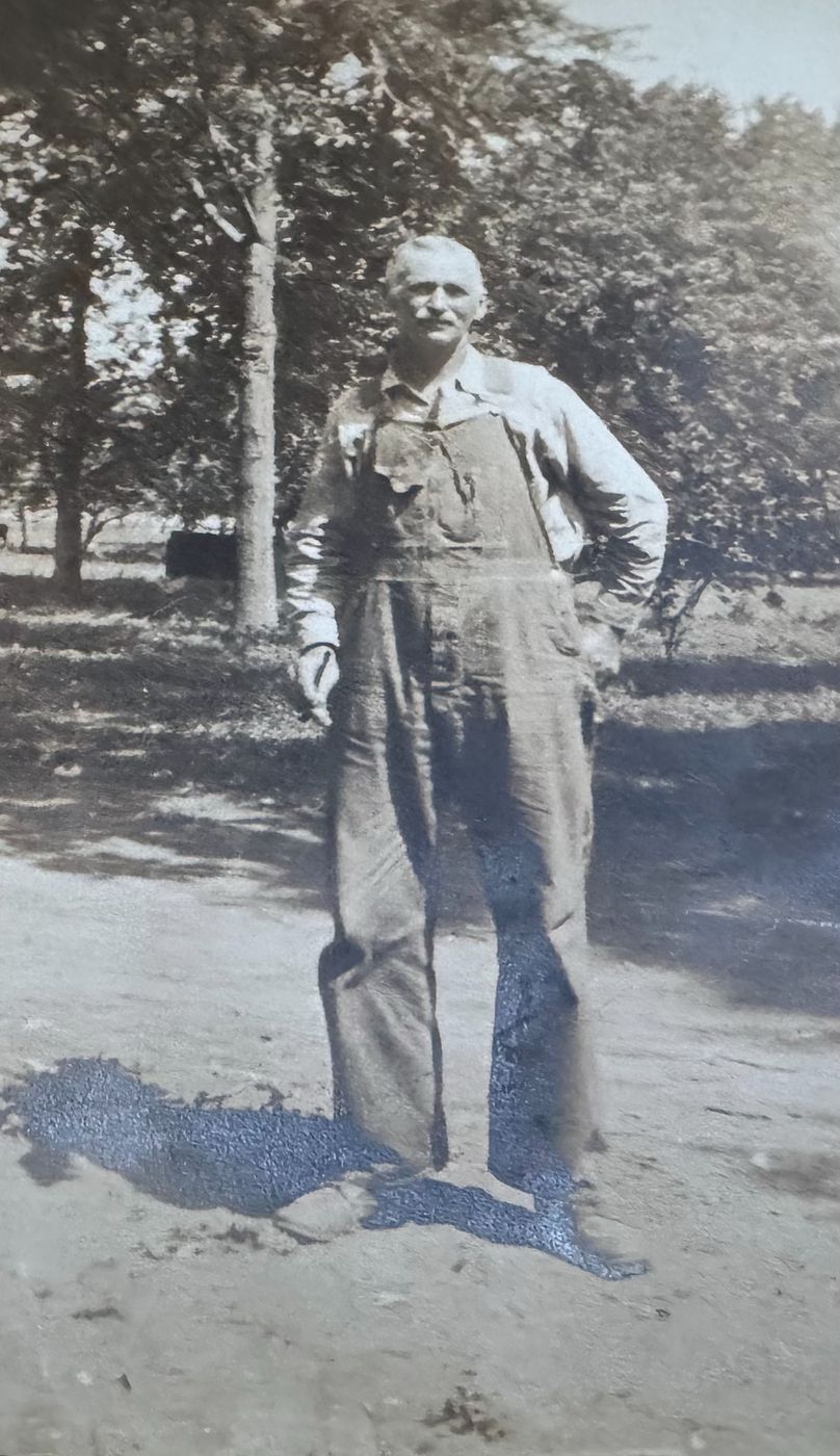 A mustached man in work shirt and overalls standing outdoors among trees.