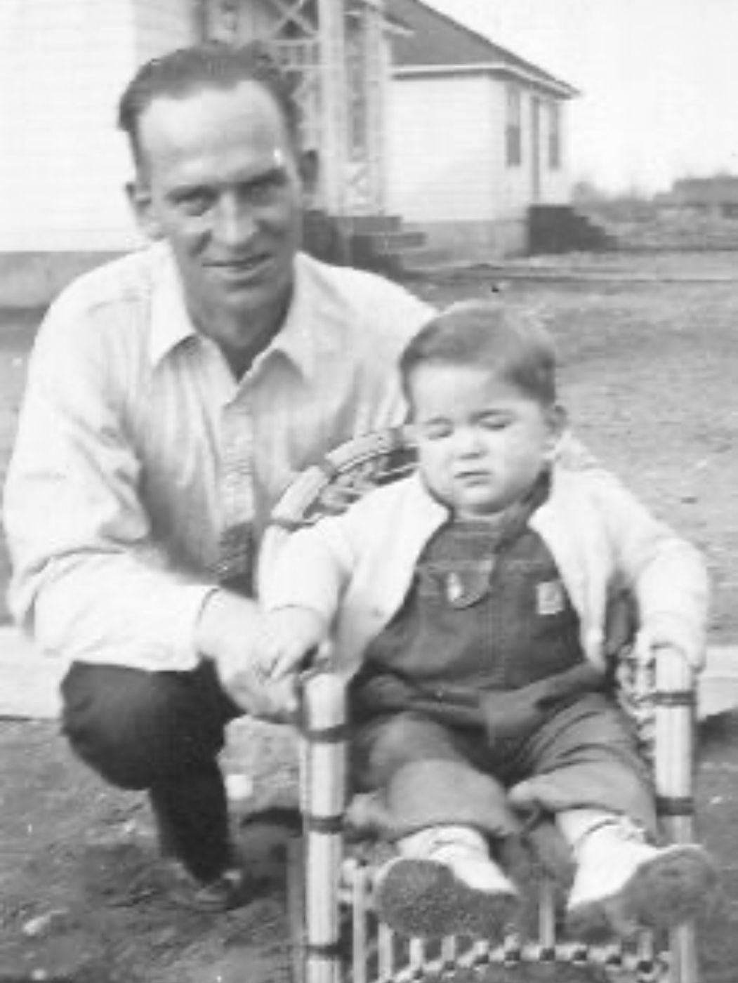 A man kneeling in a farmyard beside a small boy seated in a wicker stroller.